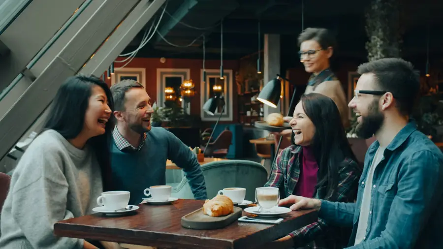 Four people laughing around a small cafe table with coffees and a croissant.