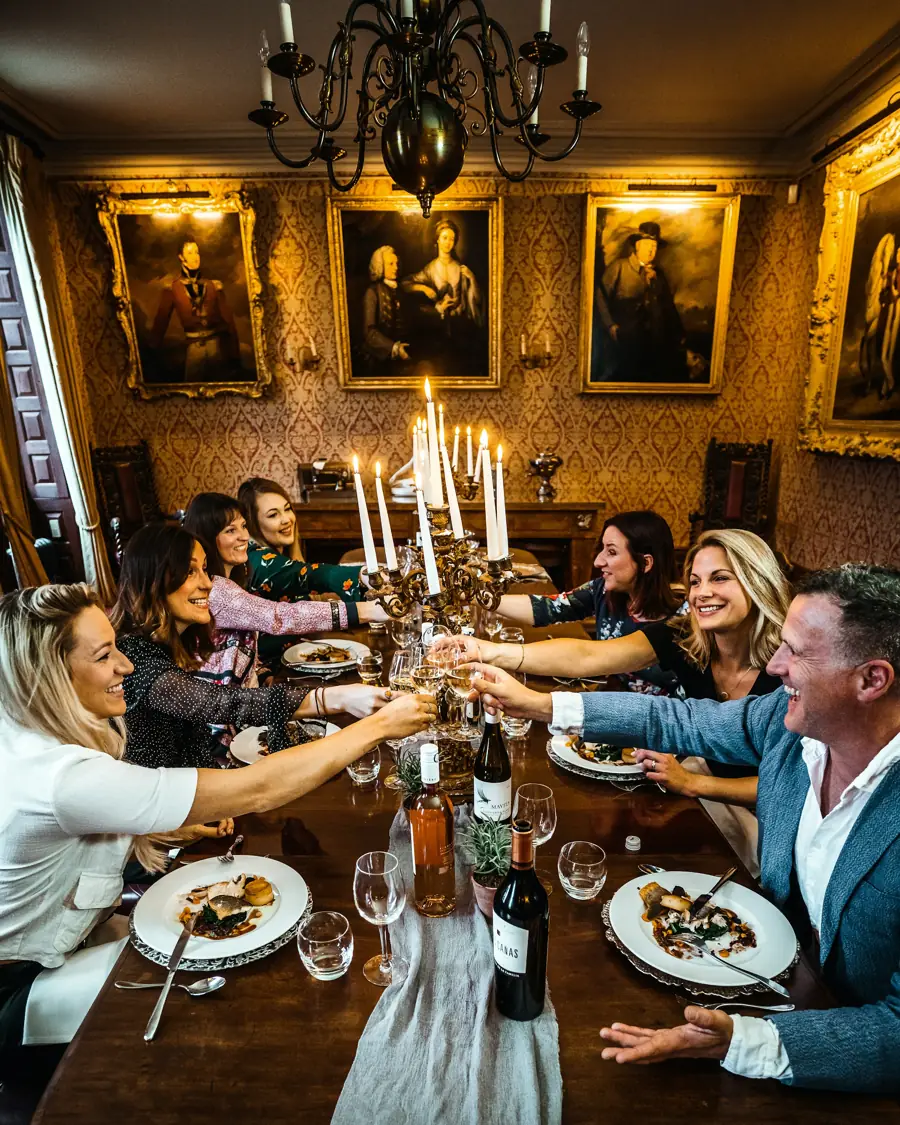 A long candlelit dinner table under a chandelier, guests raising their glasses.
