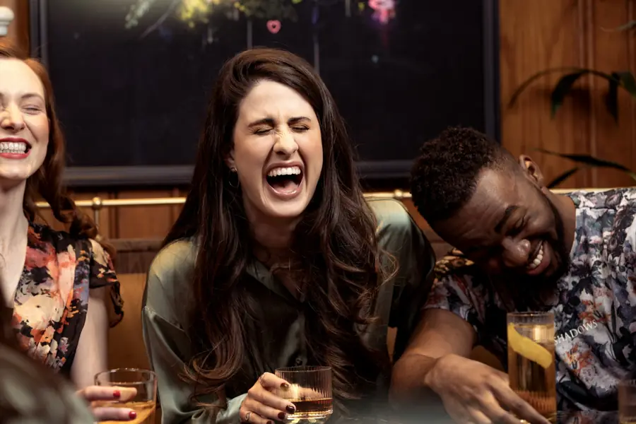 Three friends laughing hard over drinks in a warm bar.