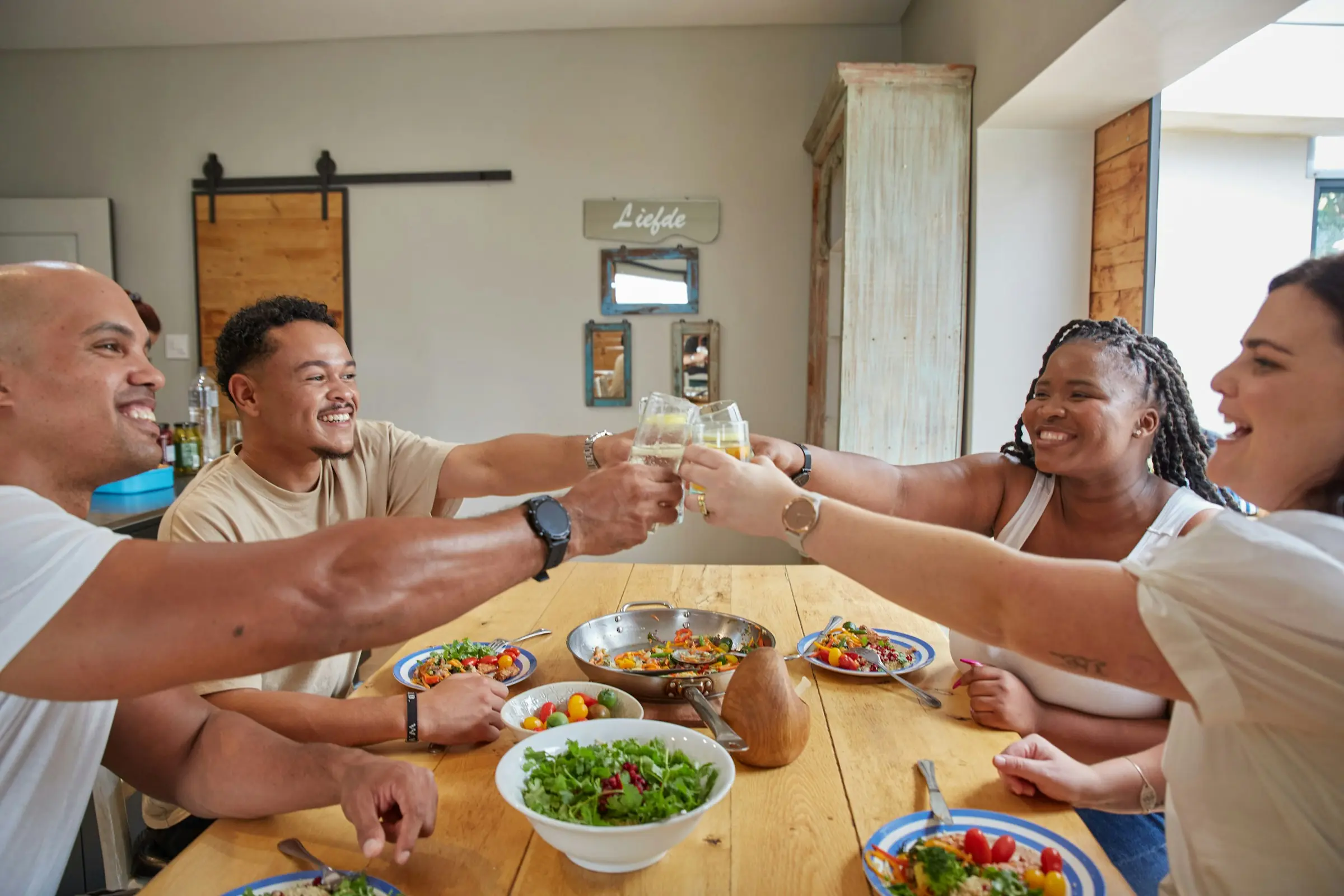 Five friends raise glasses over a long wooden table set with salads and pasta in a bright kitchen.